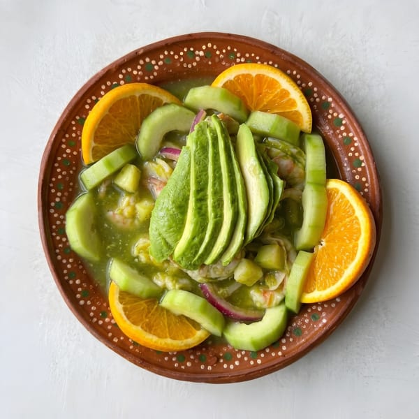 Mexican aguachile with shrimp, cucumber, avocado, red onion, and orange slices in a brown bowl.