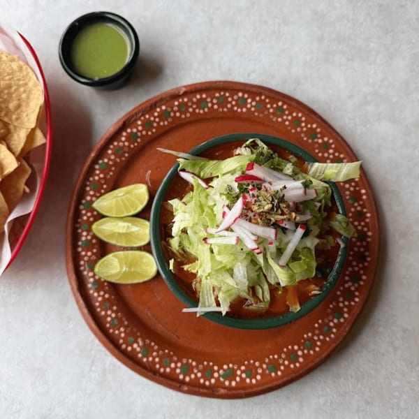 A bowl of pozole with lettuce, radishes, and spices, served with limes, chips, and green salsa.